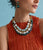 Model close-up on blue backdrop, wearing orange pleated top with Catalonia Necklace and Ethereal Pool Earrings.