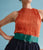 Model on blue backdrop wears orange pleated top, black skirt, and Florence Belt in Dark Emerald.