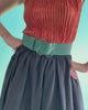 Video of model's torso on blue backdrop, in orange pleated top, black skirt, and Florence Belt in Dark Emerald.