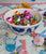 Tablescape scattered with napkins, blue glasses, flowers and Deep Bowl filled with colorful ornaments.